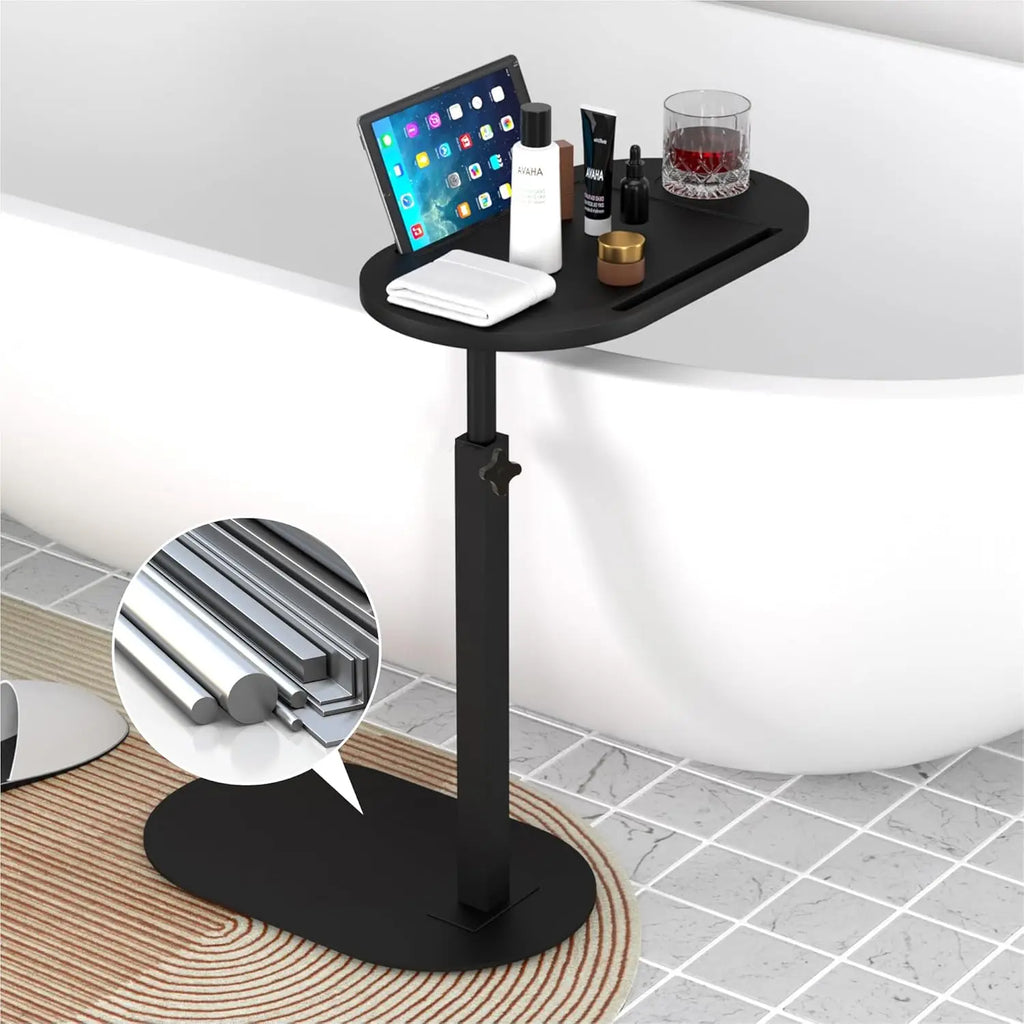 Black freestanding adjustable bathtub side tray table placed beside a tub, holding a tablet, toiletries, and a glass