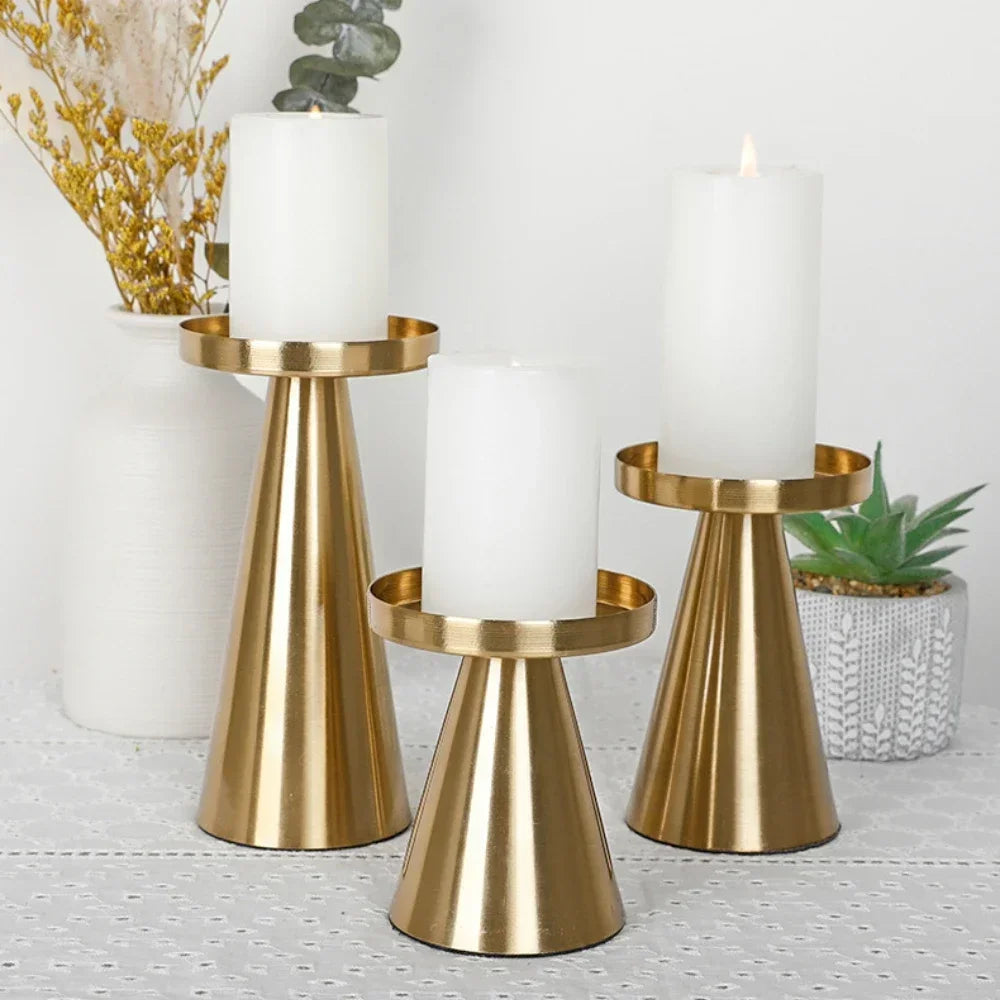 Set of three gold cone-shaped metal candle holders each with a white pillar candle, arranged on a lace tablecloth with a white vase and succulent in the background.
