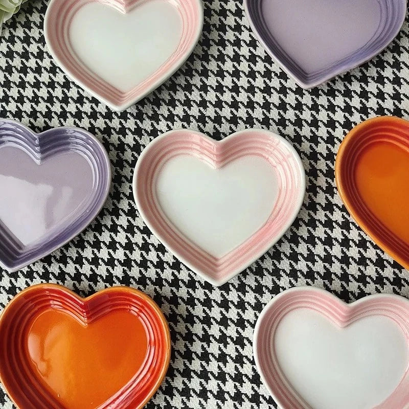 Assorted heart-shaped ceramic dishes in pink-white, purple, and orange arranged on patterned fabric