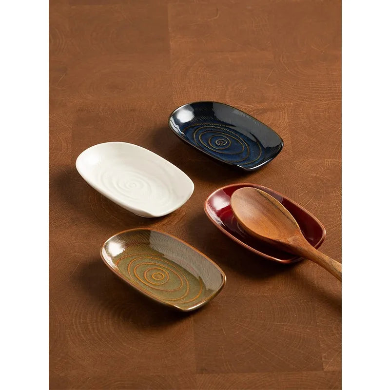 Four vintage-style oval ceramic spoon rests in white, blue, brown, and red arranged on a wood surface