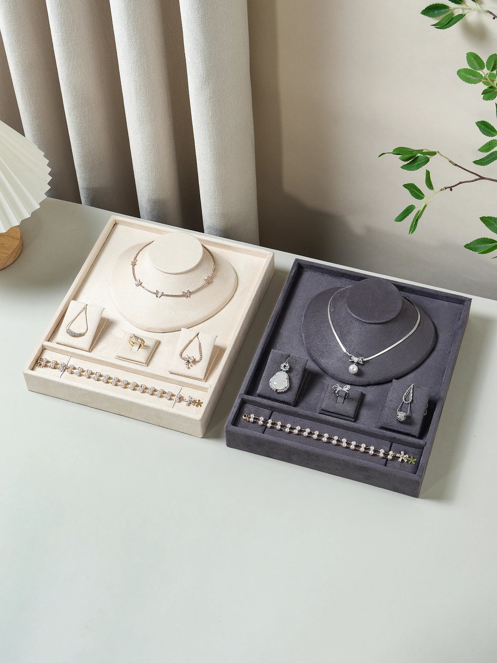 Beige and gray velvet jewelry trays holding necklaces and bracelets on a tabletop near curtains and a plant