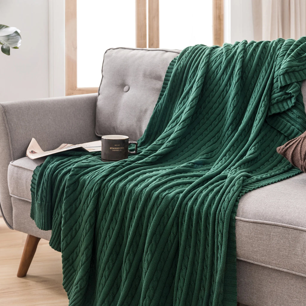 dark green cable knit cotton throw blanket draped on a sofa with a coffee mug
