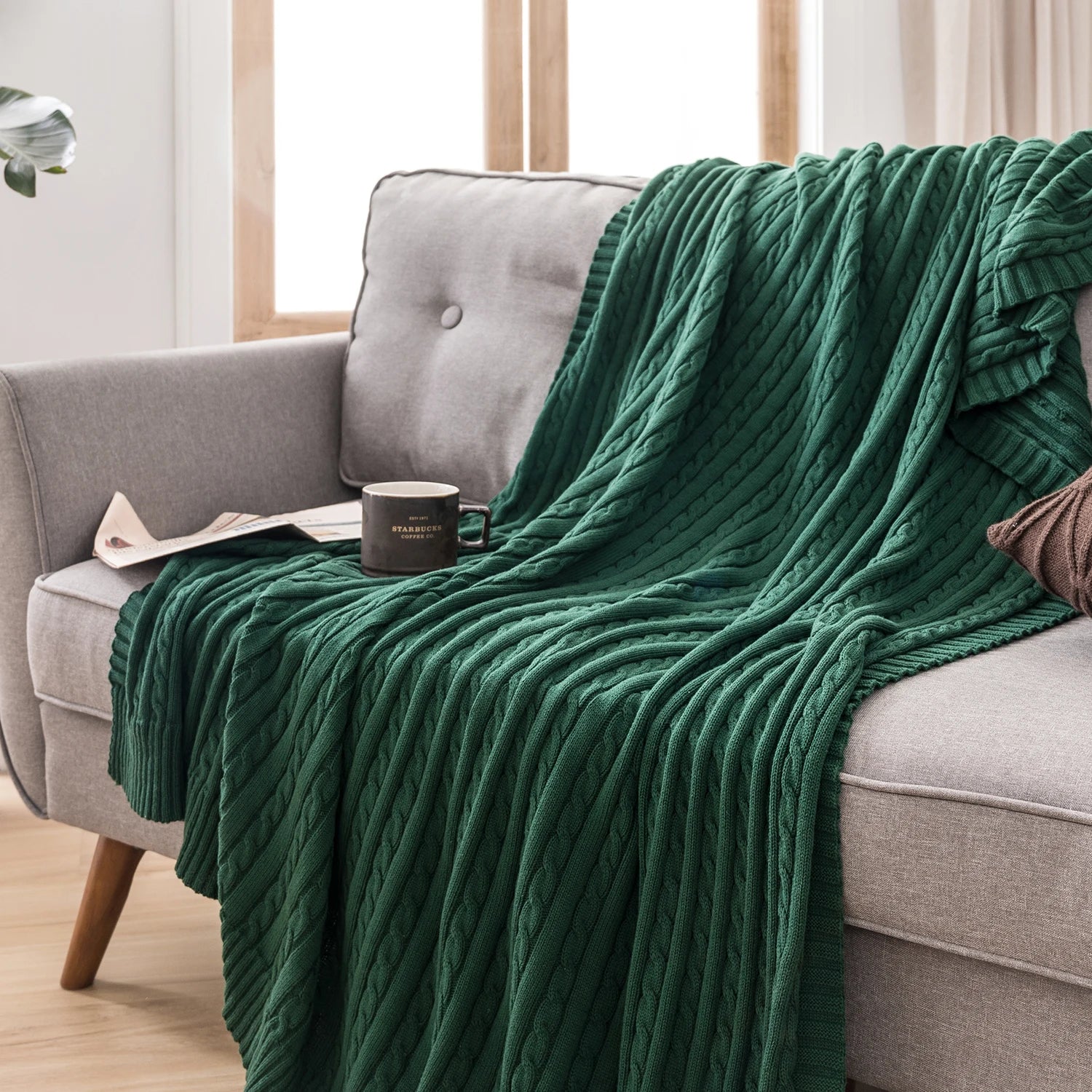 dark green cable knit cotton throw blanket draped on a sofa with a coffee mug