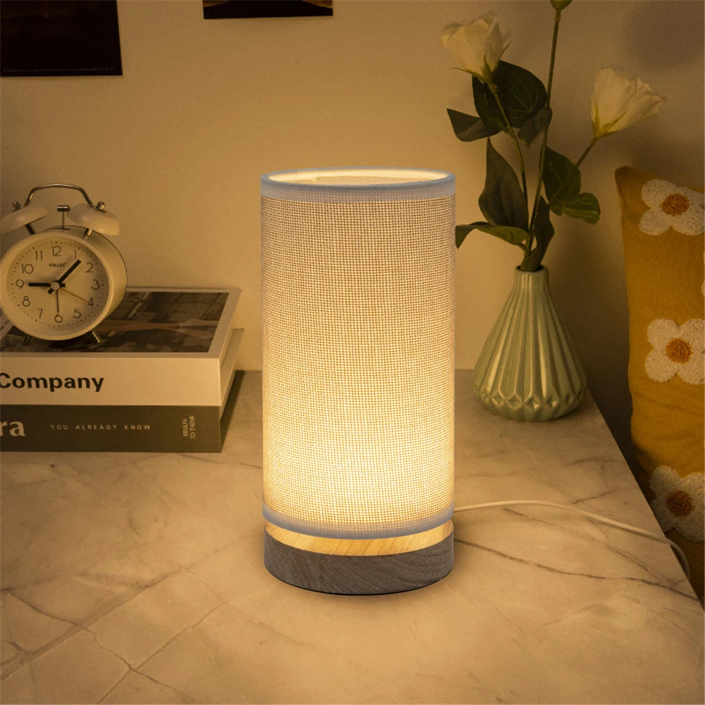 LED table lamp with cylindrical linen shade and wood base on a nightstand with books and a vase