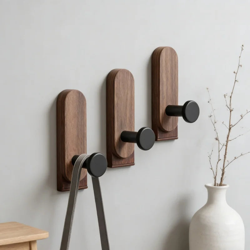 Three dark wood wall hooks with black pegs holding a bag on a neutral wall, with a minimalist vase in the background
