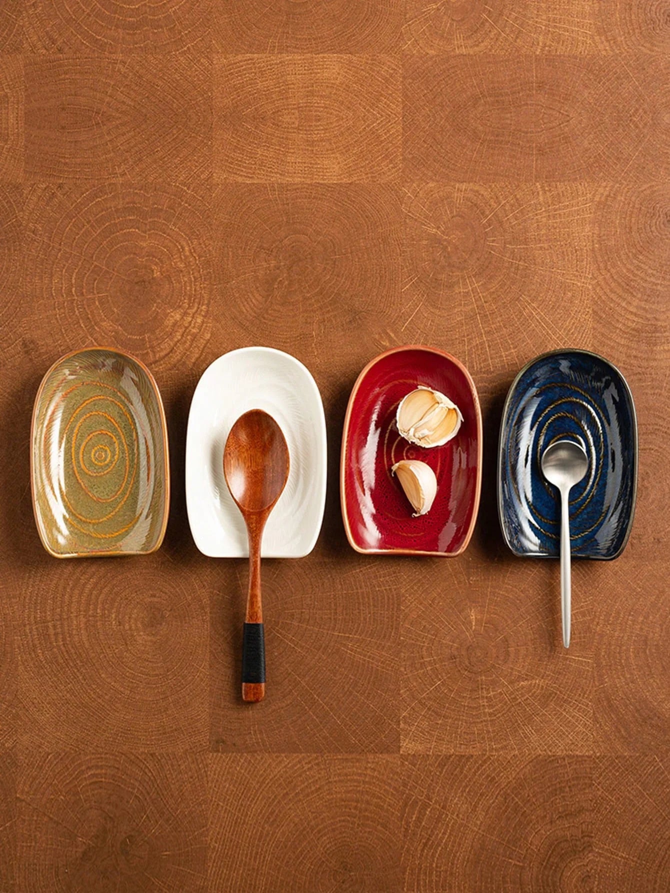Four rectangular ceramic spoon rests in gray, white, red, and blue with utensils and garlic on a wood countertop
