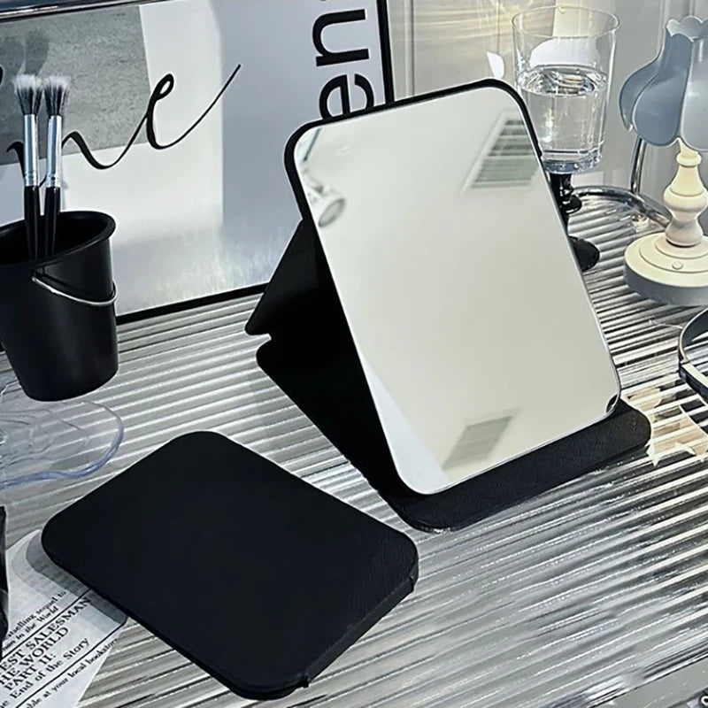Foldable black PU leather desktop makeup mirror standing on a vanity with makeup brushes and glass of water.