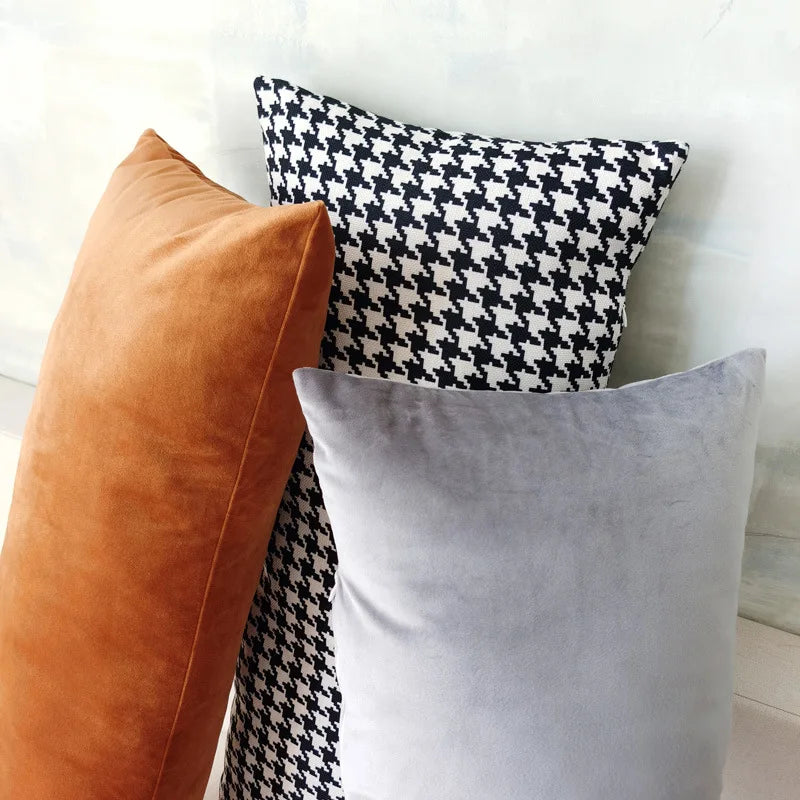 Three rectangular pillow covers in orange velvet, houndstooth, and gray velvet stacked against a wall
