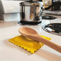 Yellow biscuit-shaped silicone spoon rest holding a wooden spoon on a white kitchen counter near a stove