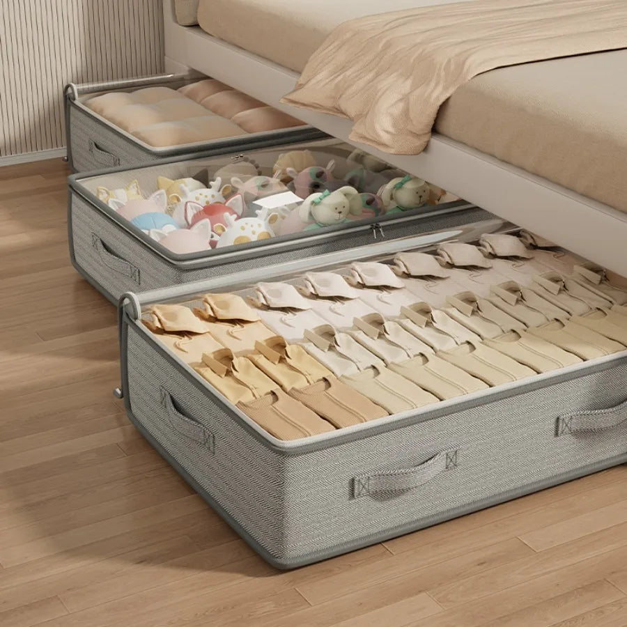 Three rectangular foldable under-bed storage bins in light green, filled with neatly folded sweaters, bedding, and soft toys, stored under a low modern bed
