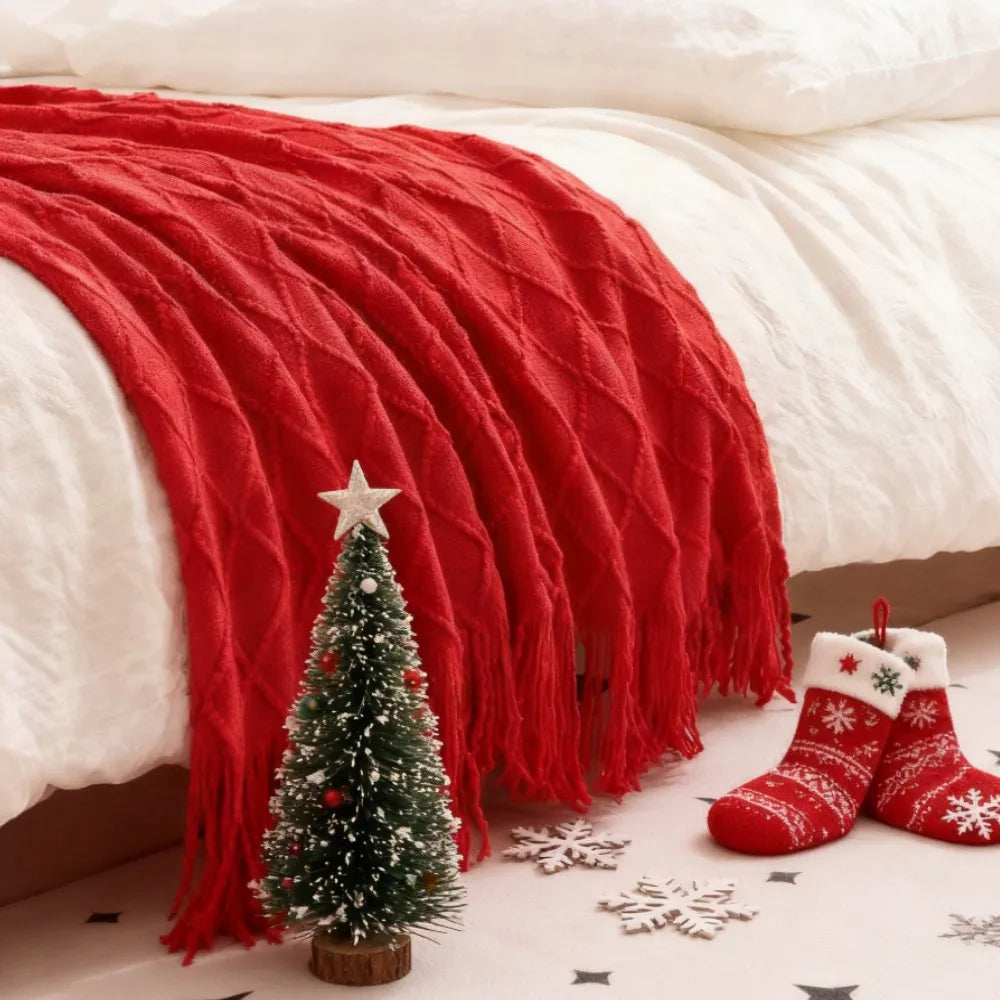Red knit throw blanket with fringe on a bed, next to Christmas decorations and mini tree