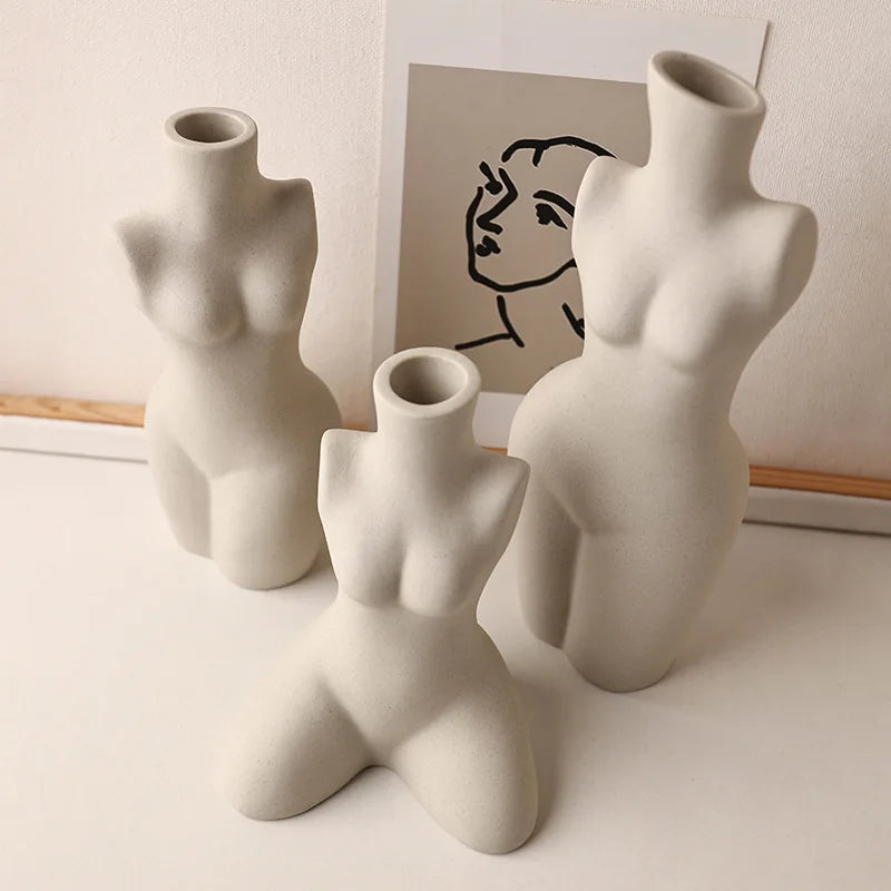 Three light ceramic body form vases grouped on a white surface with minimalist art in the background