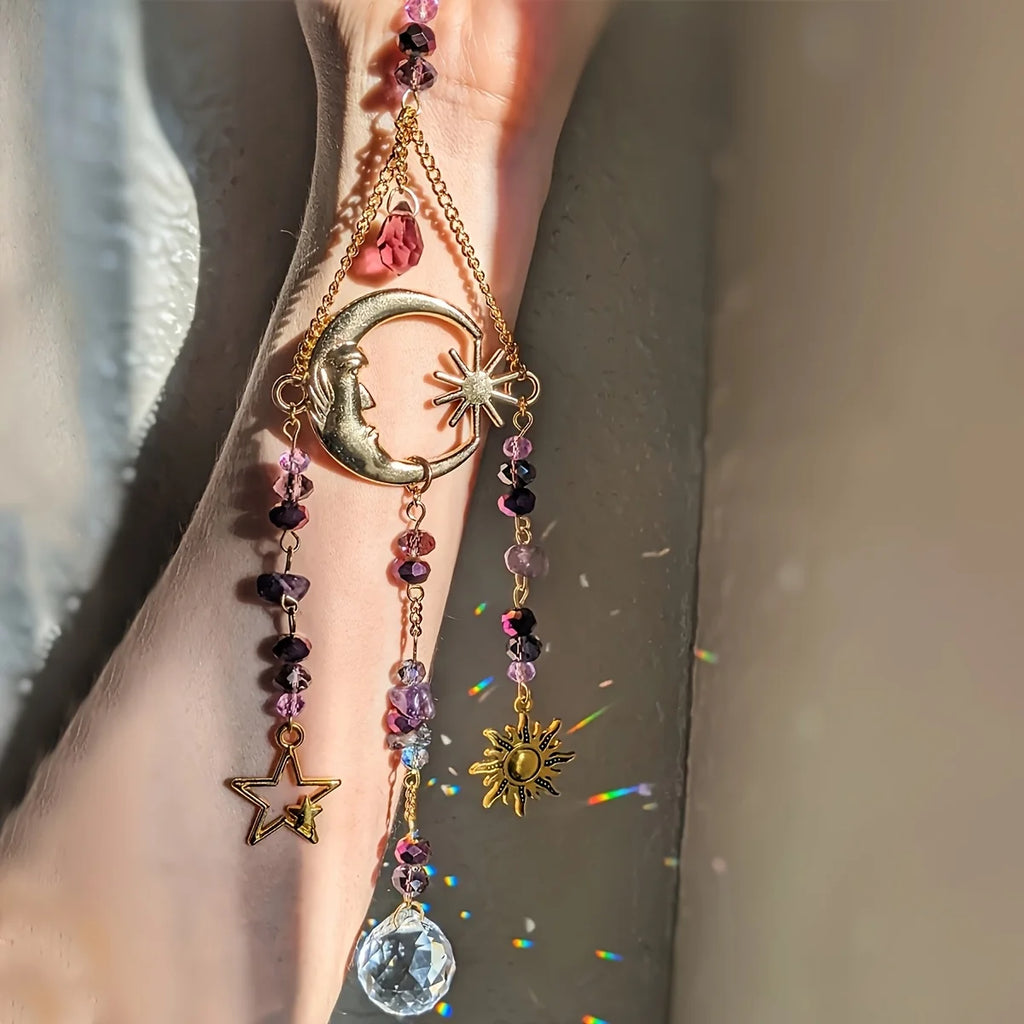Gold celestial crystal suncatcher with moon, sun, and star charms hanging in sunlight and creating subtle rainbow sparkles.