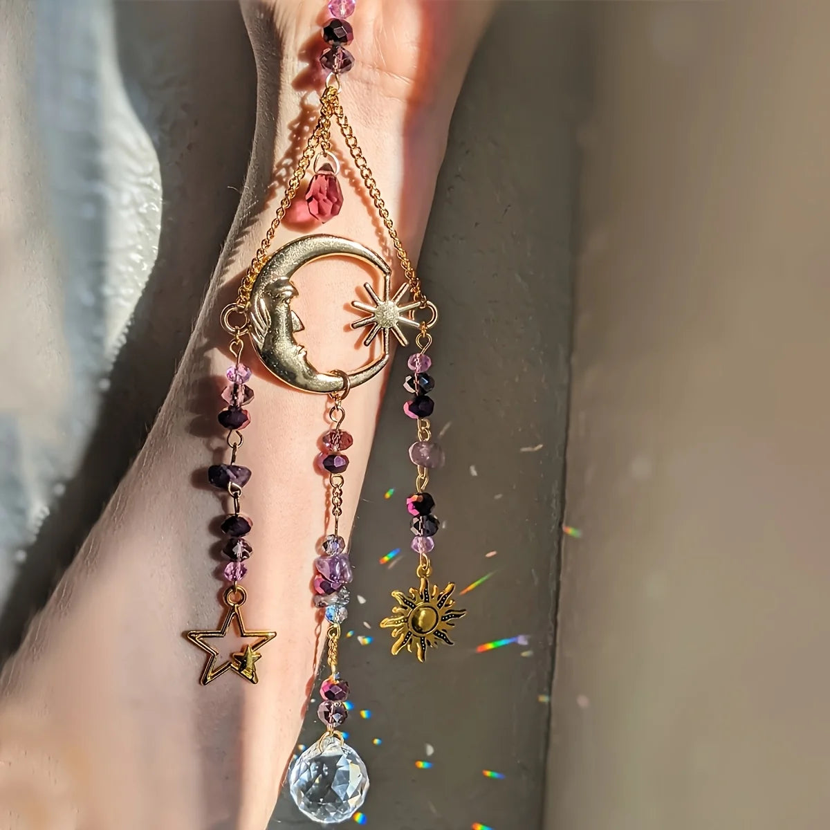 Gold celestial crystal suncatcher with moon, sun, and star charms hanging in sunlight and creating subtle rainbow sparkles.