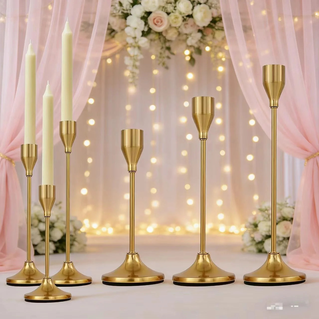 Three gold metal candle holder sets arranged on a wedding table with taper candles, set against a pink draped floral backdrop