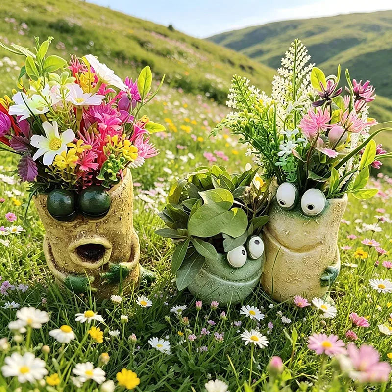 Three frog-shaped resin planters with plants outdoors in a wildflower field