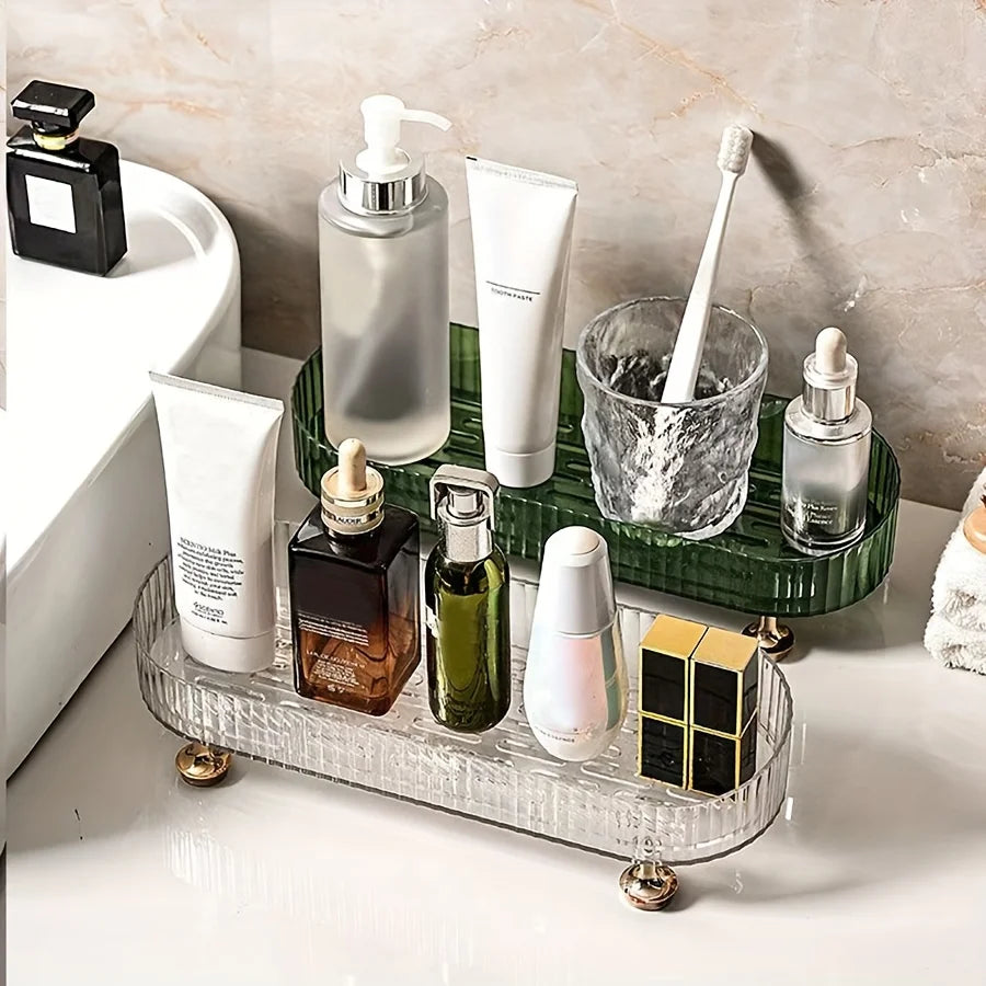 Clear and green acrylic vanity trays with gold feet, each organizing an assortment of skincare bottles and toiletries on a bathroom countertop