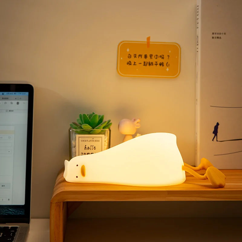 Lying duck silicone night light glowing on a wooden desk beside a succulent and books