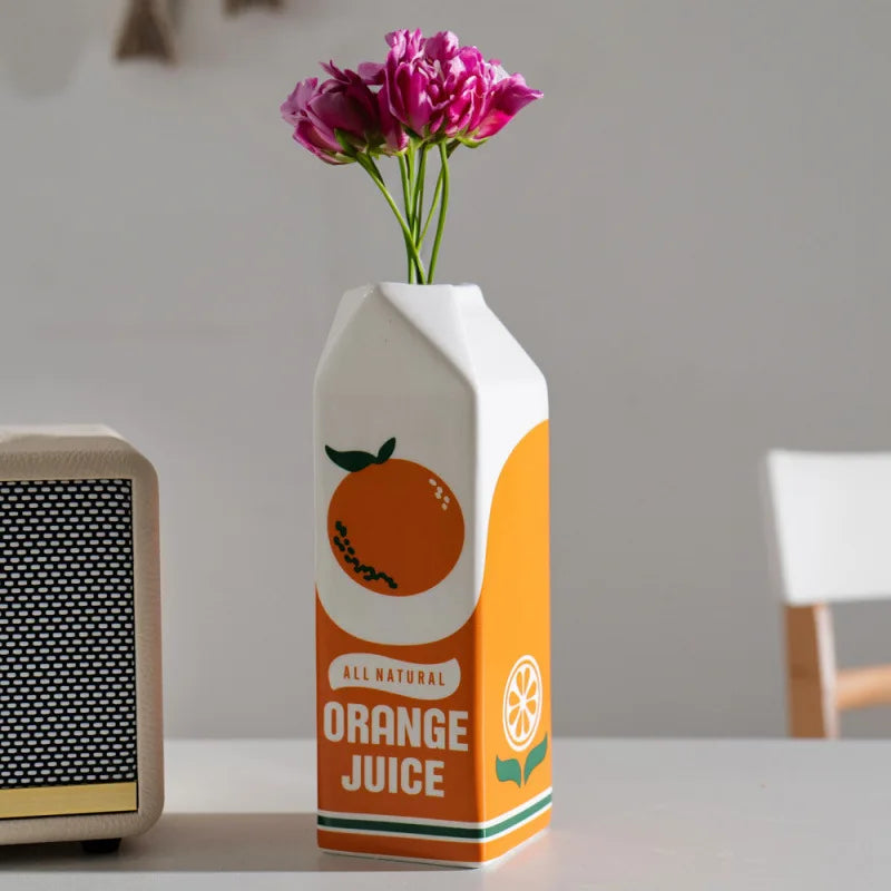 Ceramic orange juice carton vase with pink flowers on a tabletop in a modern home setting
