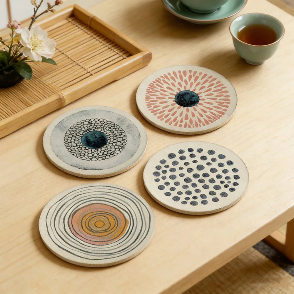 Four colorful round art coasters arranged on a light wood coffee table next to tea cups