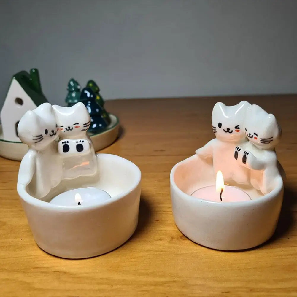 Two cat tealight candle holders on a table, each holding a lit tealight, with decorative trees in the background
