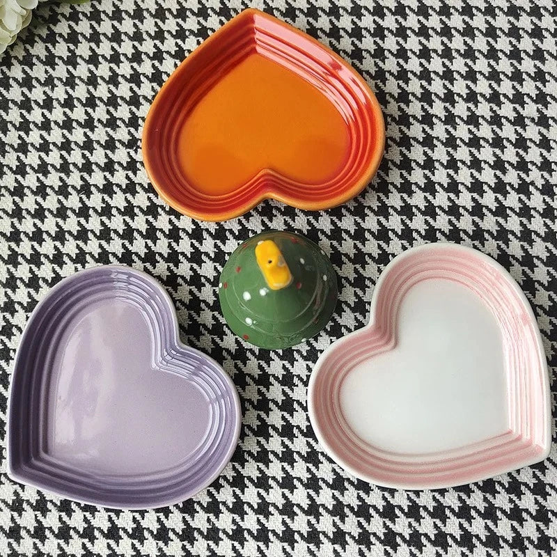 Three heart-shaped ceramic dishes in orange, purple, and pink-white on a houndstooth patterned surface