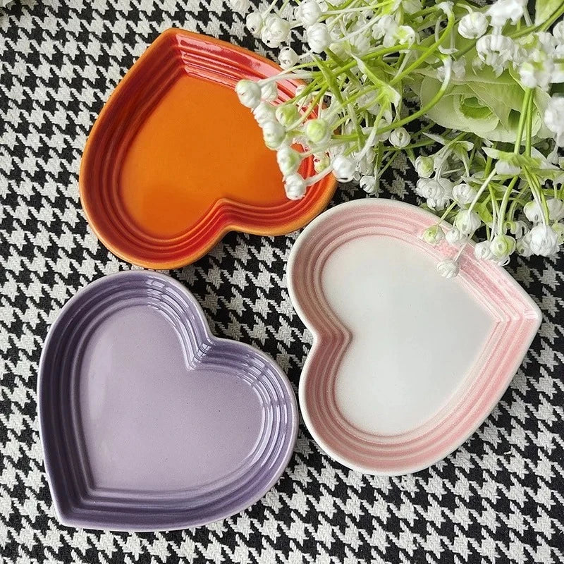 Set of heart-shaped ceramic dishes in orange, purple, and pink-white with white flowers nearby
