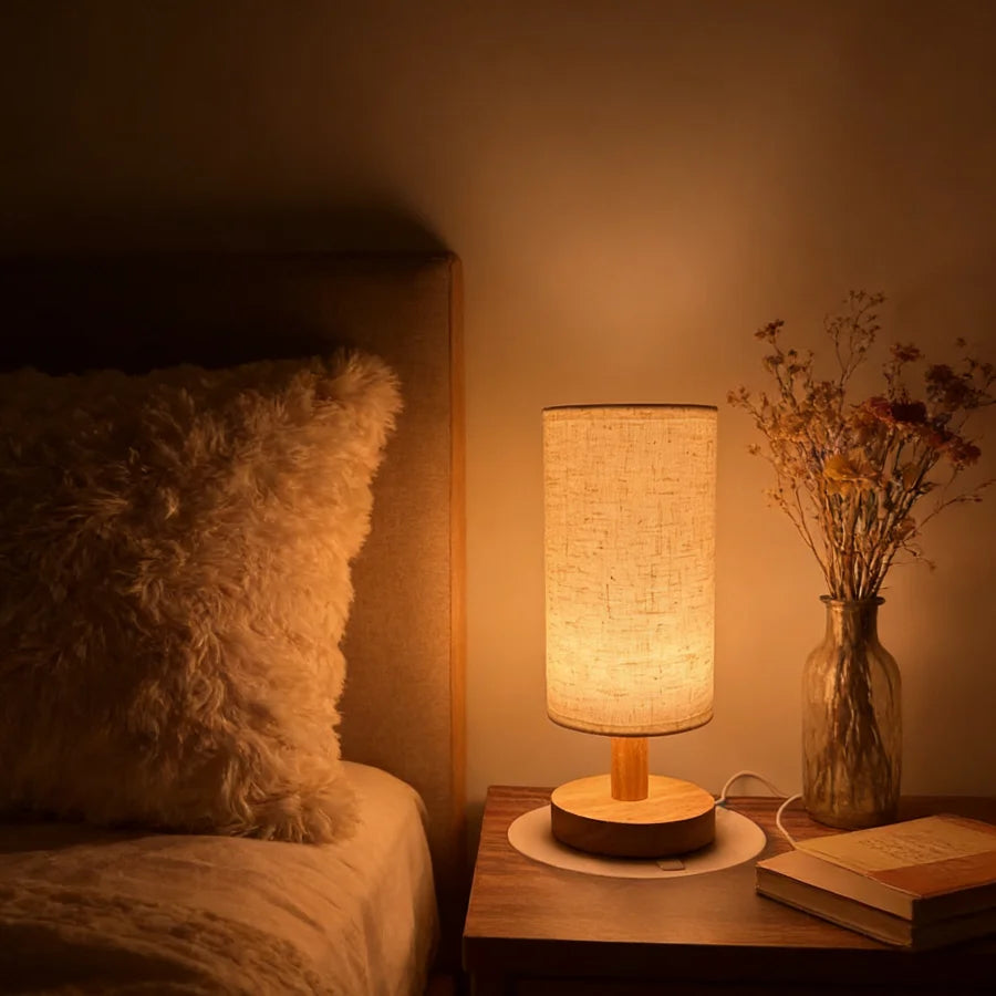 Wooden bedside table lamp with linen shade casting a warm glow on a nightstand beside a cozy bed with a fluffy pillow and dried flowers in a vase