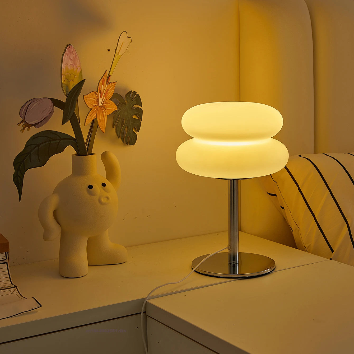 Cloud-shaped table lamp with warm yellow light on a nightstand beside a cream vase and bed