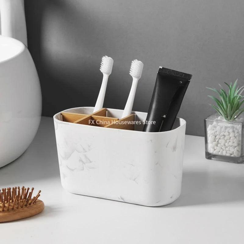 White marble toothbrush holder with bamboo dividers holding two toothbrushes and a toothpaste tube, placed near a sink and a plant