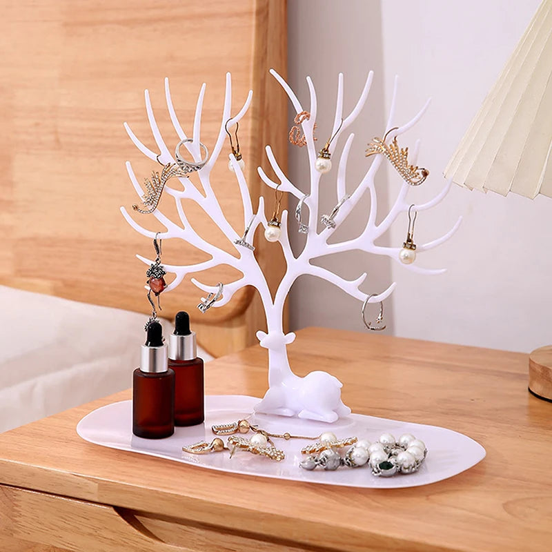 White reindeer tree jewelry organizer with tray on a wooden nightstand, storing earrings, rings, and bottles