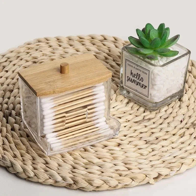 Clear cotton swab holder with bamboo lid on woven mat next to small succulent in a glass container