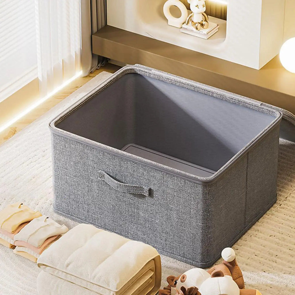 Open grey fabric storage box on a rug with folded blankets and toys nearby