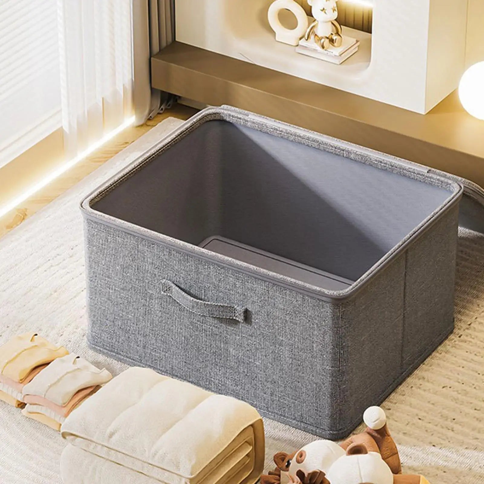 Open grey fabric storage box on a rug with folded blankets and toys nearby