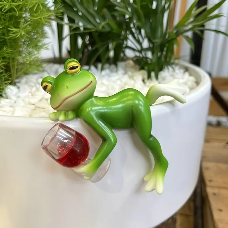Cartoon resin frog figurine with a wine glass, resting on the edge of a planter with white pebbles.
