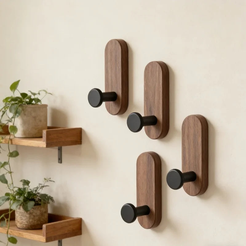 Four dark wood self-adhesive wall hooks with black pegs on a light wall in an entryway, next to wood shelves and potted plants