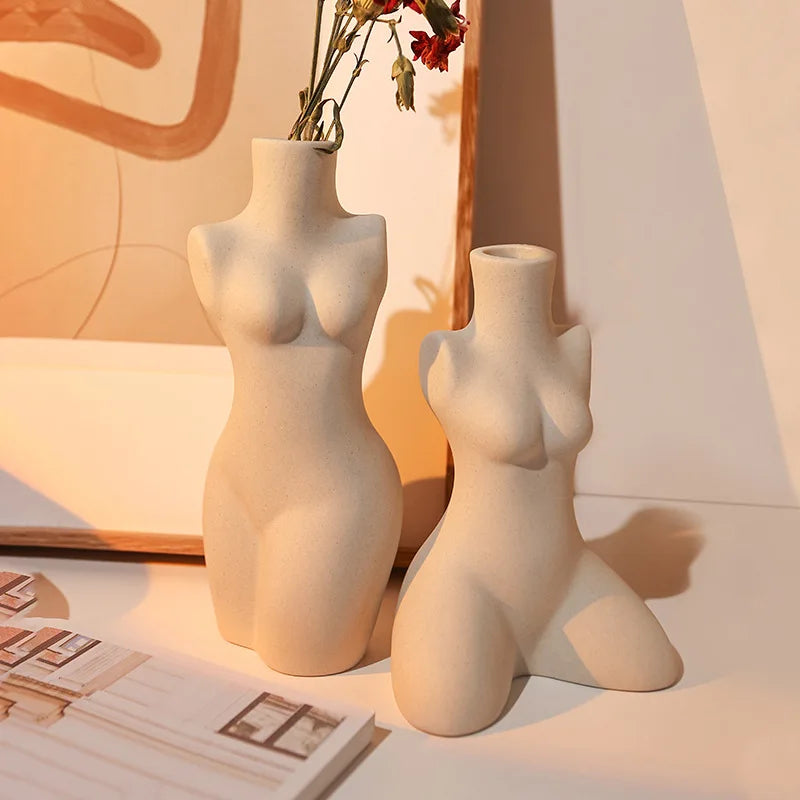 Two beige ceramic torso vases with red flowers, styled on a desk beside artwork and lamp
