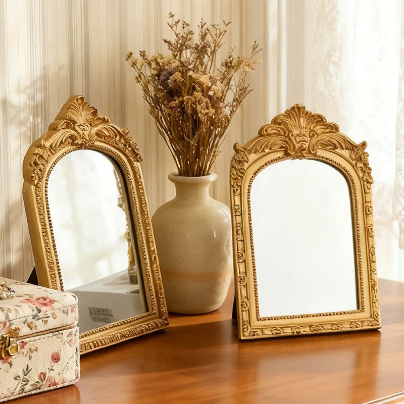 Two gold arched carved mirrors on a wooden dresser near a vase of dried flowers