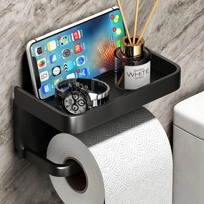 Wall-mounted black aluminum toilet paper holder with top storage shelf holding a phone, watch, and diffuser, mounted on a bathroom wall.