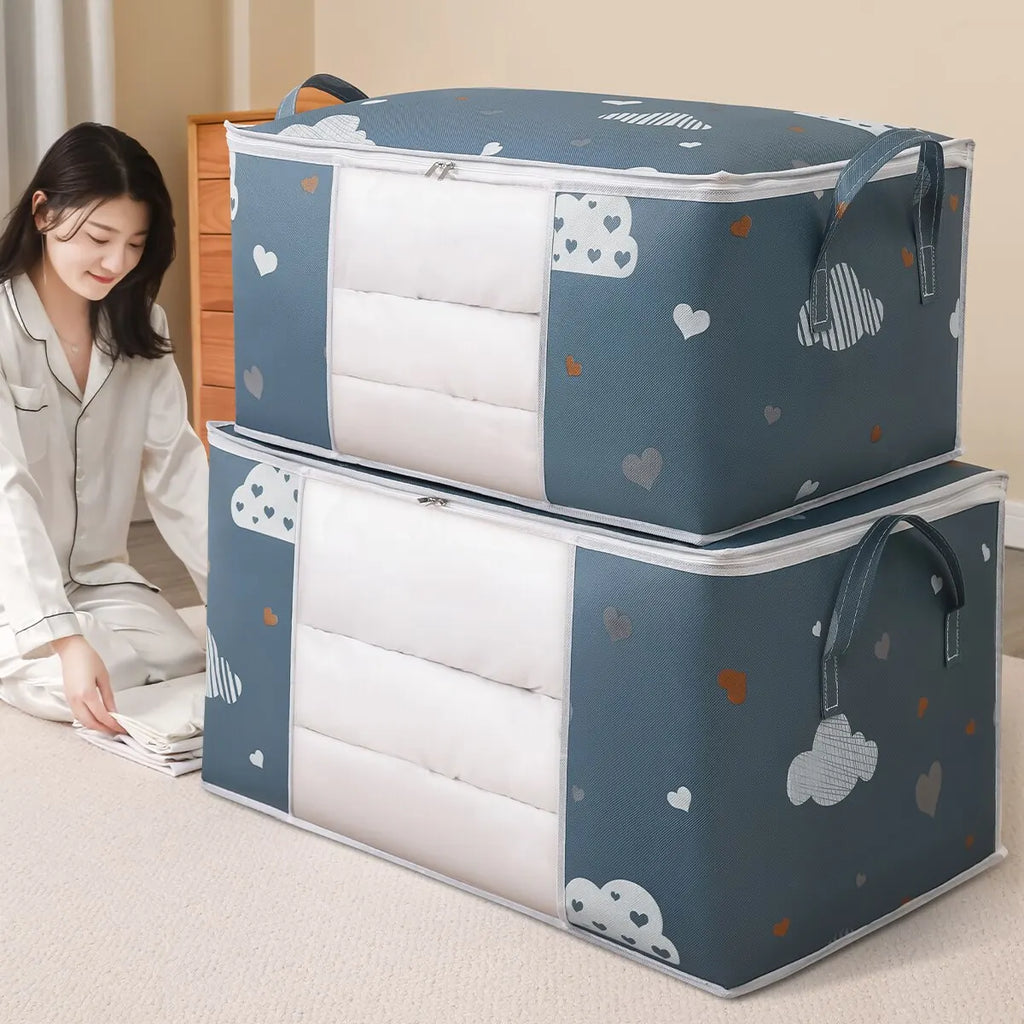 Two large blue cloud print foldable storage bags with reinforced handles stacked in a bedroom, one being filled with bedding by a person.