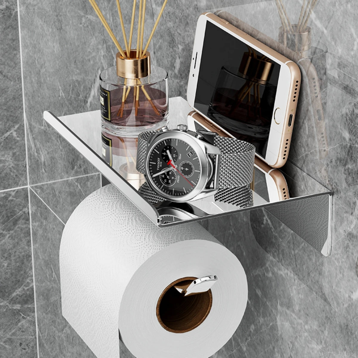 Stainless steel toilet paper holder with shelf, shown holding a watch, phone, and diffuser bottle mounted on a bathroom wall