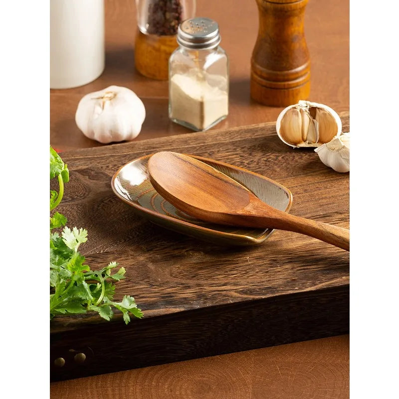 Close-up of a green-brown ceramic spoon rest holding a wooden spoon on a wooden board with garlic and herbs nearby