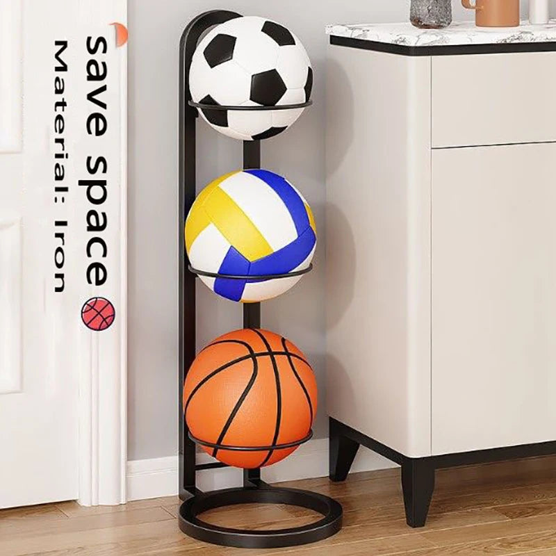 Black metal ball storage rack with three tiers holding various sports balls, placed next to a white cabinet against a wall.