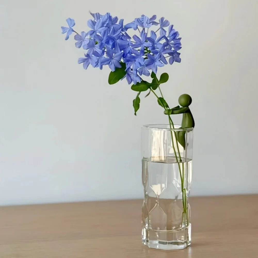 Single dark green plant stem holder clipped to a glass vase with blue flowers on a wood surface