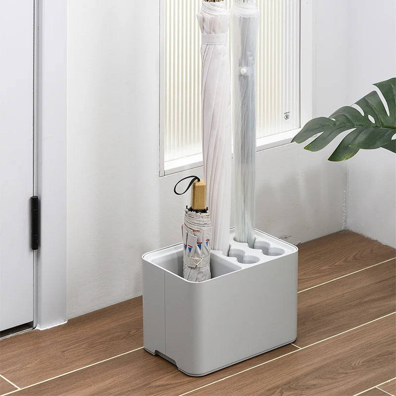 Modern plastic umbrella stand in light grey, placed in a corner entryway holding long umbrellas