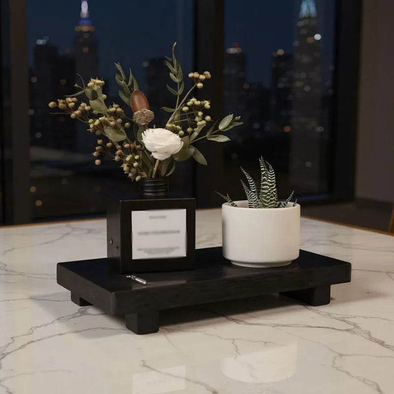 Black wooden display tray on marble table with small vase and potted plant, night city in background
