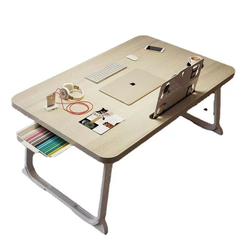 Beige foldable bedside table with a laptop, headphones, books, and integrated side drawer holding colored pens