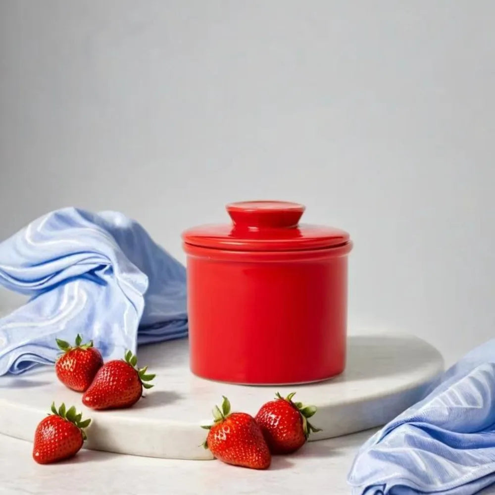 Red ceramic French butter crock with lid displayed on a white platter with strawberries