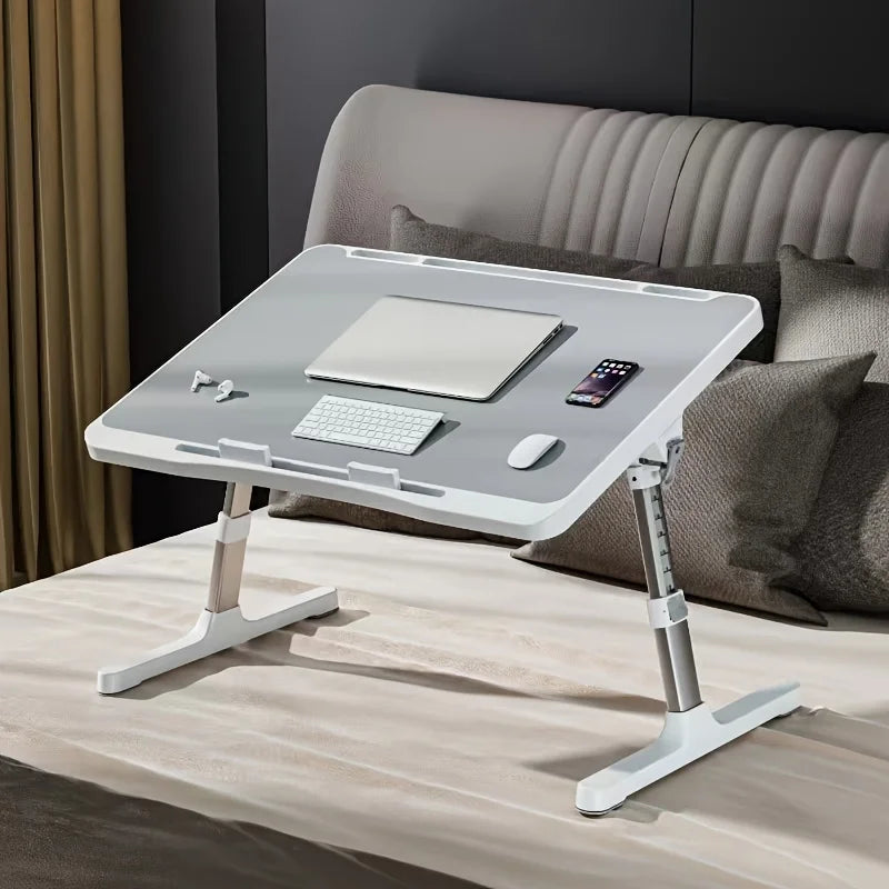Adjustable gray lap desk on a bed arranged with laptop, keyboard, mouse, and phone