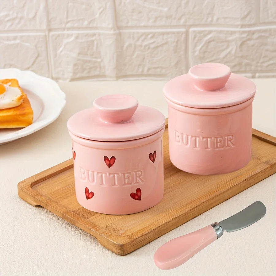 Two pink ceramic butter crocks with embossed 'BUTTER' label, one with heart accents, set on a wooden tray; matching pink-handled butter knife included.